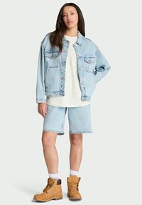 Jaqueta de ganga azul clara com bolsos no peito, t-shirt branca, calções azuis claros e botas castanhas com solas cinzentas. O modelo está em frente a um fundo simples.