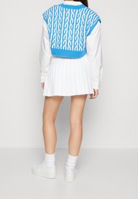 Blå stickad ärmlös tröja med ett vitt kabelmönster, kombinerad med en vit veckad kjol och vita sneakers. Bakifrån.