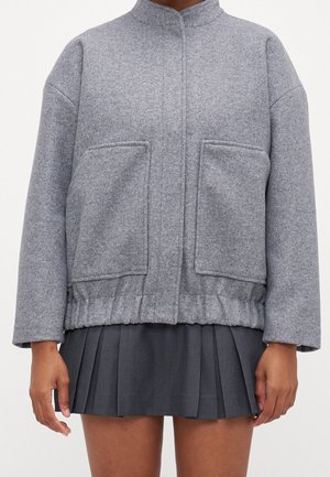 Veste en laine grise avec poches avant portée sur une mini-jupe plissée foncée, visible des épaules jusqu'à mi-cuisse.