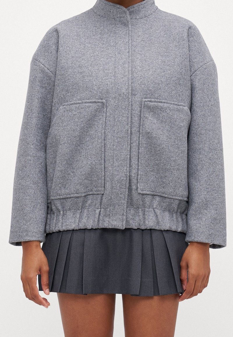 Veste en laine grise avec poches avant portée sur une mini-jupe plissée foncée, visible des épaules jusqu'à mi-cuisse.