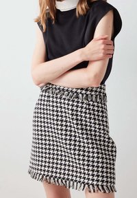 Falda en blanco y negro de patrón houndstooth con dobladillo desflecado, hecha de tela tejida. Combinada con una blusa negra con un alto cuello blanco.