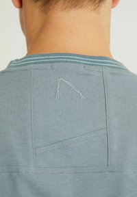 Hellblaues T-Shirt mit einem gerippten Halsausschnitt mit blauen Streifen, einer gesteppten Naht und einer kleinen Tasche auf der Rückseite. Weiche Stoffstruktur.