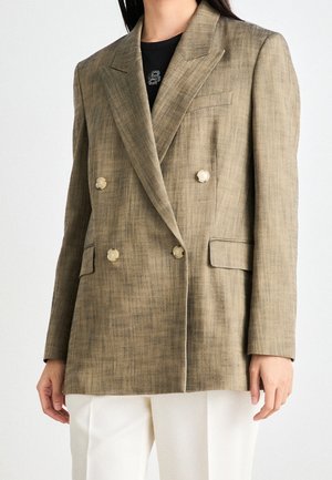 Žena v béžovém dvouřadovém saku se čtyřmi knoflíky přes černý top a bílé kalhoty, stojící před jednobarevným pozadím.