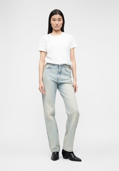 Νεαρή γυναίκα όρθια, φοράει λευκό μπλουζάκι, ανοιχτό μπλε τζιν με ψηλή μέση και μαύρα ψηλοτάκουνα μπότες μέχρι τον αστράγαλο σε απλό φόντο.