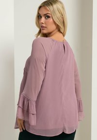 Blusa lavanda semi-trasparente con maniche a campana, schiena a oblò e dettagli in perline lungo i polsini. Tessuto morbido e testurizzato.