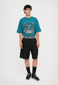 Zöldes kék, oversized t-shirt grafikus mintával, fekete térdhez érő sorttal és fekete sportcipővel párosítva. Kontrasztos derékpántja van.