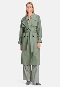 Light green trench coat with a belted waist, large lapels, and double-breasted buttons. Paired with patterned wide-leg pants and pointy shoes.