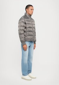 Jachetă puffer gri cu un model în diamant, guler înalt, închideri cu fermoar, asortată cu blugi drepti de culoare albastră deschisă și pantofi sport albi.
