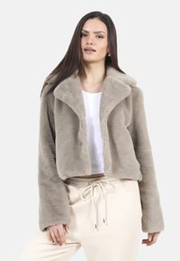 Przycięta, beżowa kurtka z futra syntetycznego z szerokim kołnierzem i długimi rękawami. Nosi się ją na białej bluzce, zestawiona z jasnobeżowymi spodniami.