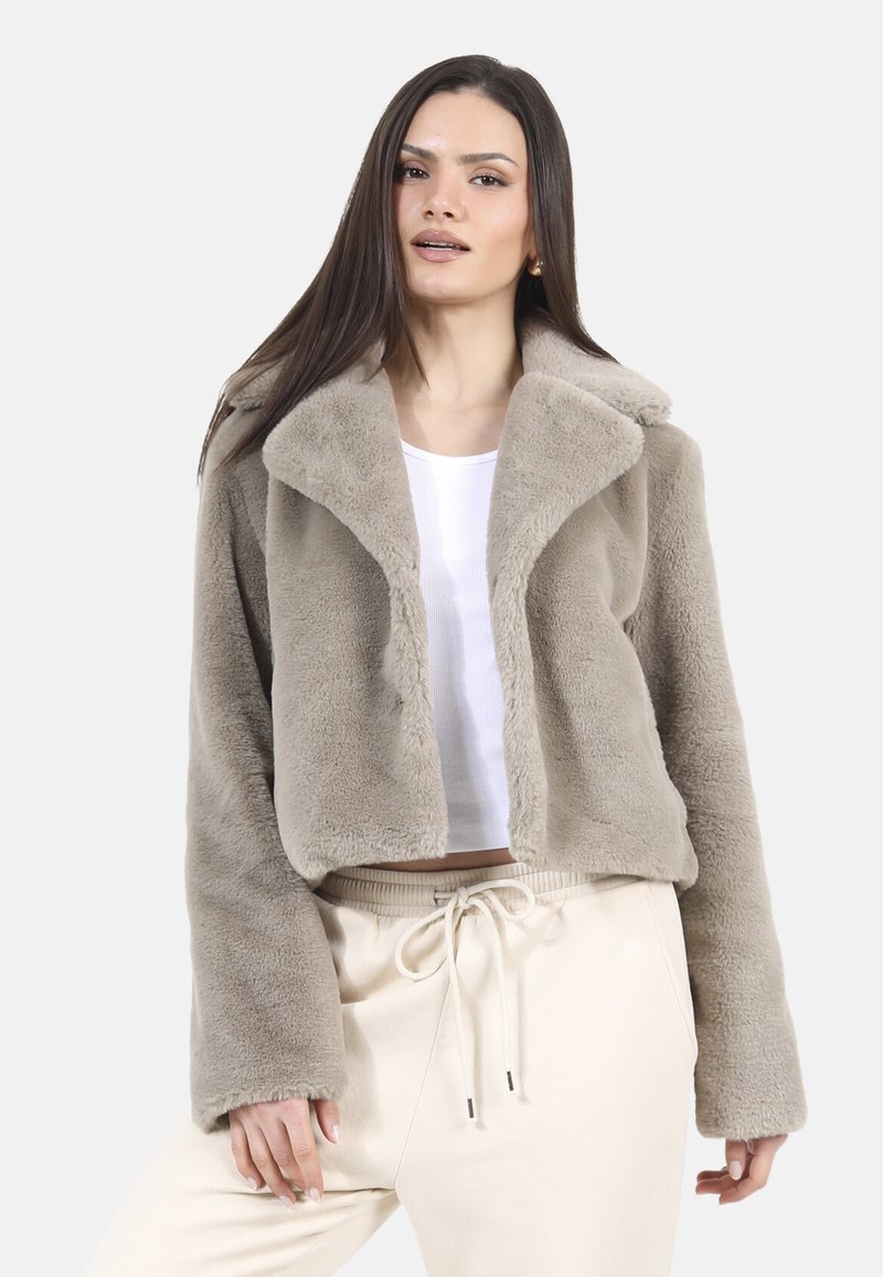 Przycięta, beżowa kurtka z futra syntetycznego z szerokim kołnierzem i długimi rękawami. Nosi się ją na białej bluzce, zestawiona z jasnobeżowymi spodniami.