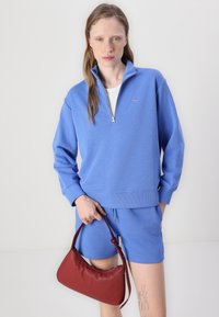 Svetlomodrý zippered sveter vyrobený z mäkkej látky s rebrovaným spodkom, skombinovaný s hodiacimi šortkami, drží malú červenú koženú kabelku.
