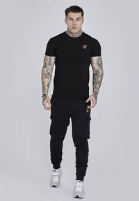 Czarna koszulka z okrągłym dekoltem i haftowanym logo, zestawiona z czarnymi spodniami typu cargo z kieszeniami. Noszona z białymi tenisówkami.