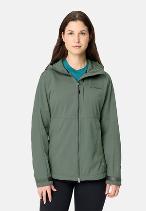 Donna che indossa una giacca verde con cappuccio e zip sopra una maglietta verde acqua, in piedi di fronte a uno sfondo semplice.