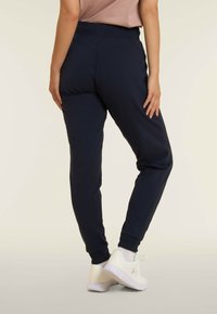 Joggers blu navy con vita a coste e orli delle gambe affusolati, realizzati in un tessuto liscio. Indossati con scarpe da ginnastica bianche.