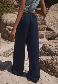 Wide-leg navy trousers feature a soft, lightweight fabric and an elastic waistband, set against a sandy beach with textured rocks.