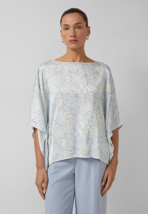 Kobieta w luźnej, jasnoniebieskiej bluzce w kwiatowy wzór z szerokimi rękawami i dopasowanych jasnoniebieskich spodniach, stojąca na jednolitym tle.