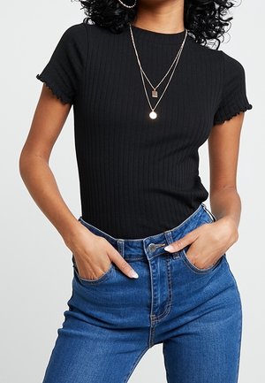 Vrouw die een zwart geribbeld shirt met korte mouwen en geplooide randen draagt, met gelaagde gouden kettingen en een hoge taille blauwe spijkerbroek, met haar handen in de zakken.