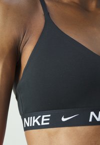 Crni sportski grudnjak s elegantnim dizajnom, koji ima podesive naramenice i široki pojas s bijelim tekstom "NIKE". Glatka tekstura tkanine.