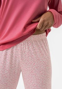 Une personne portant un haut à manches longues rose le soulève légèrement pour montrer la taille élastique d'un pantalon blanc avec un petit motif floral rose.
