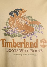T-shirt graphique beige présentant des bottes Timberland illustrées en brun, entourées de plantes vertes et violettes. Le texte indique "Bottes Timberland avec Racines."