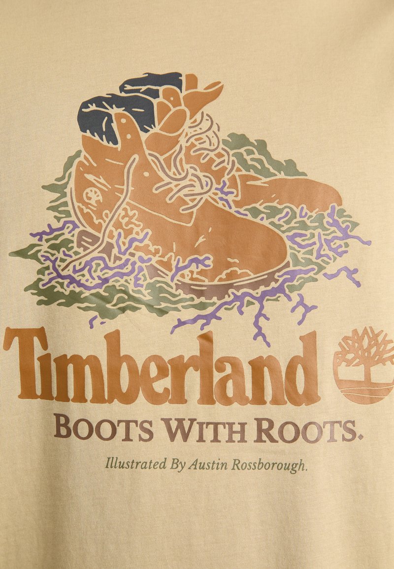 Beige Grafik-T-Shirt mit illustrierten braunen Timberland-Stiefeln, umgeben von grünen und lila Pflanzen. Der Text lautet "Timberland-Stiefel mit Wurzeln."