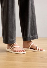 Strappy sandals in light beige with a flat sole, featuring two crossed straps and a toe loop, paired with gray wide-leg pants.