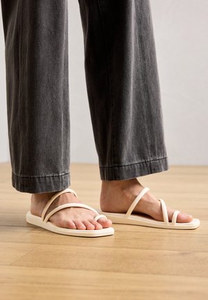 Lichtbeige sandalen met banden en een platte zool, uitgerust met twee gekruiste banden en een teenlus, gecombineerd met grijze wijduitlopende broek.