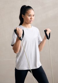 Pasipriešinimo juostos su ergonomi�škomis juodomis rankenomis, kurias laiko žmogus šviesiai pilkoje marškinėliuose ir juodose tamprėse, prieš neutralų foną.