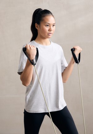 Bande elastiche con manici ergonomici neri impugnate da una persona che indossa una maglietta grigia chiara e leggings neri, sullo sfondo neutro.