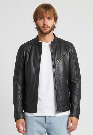 Hombre con chaqueta de cuero negra con cremallera sobre una camiseta blanca y vaqueros azul claro, de pie frente a un fondo liso.
