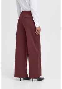 Wijdgevallen broek in bordeauxkleurige stof met een elastische tailleband. Heeft een achterzak en wordt gedragen met zwarte hoge hakken.