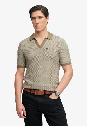 Mann trägt kurzärmeliges, strukturiertes beiges Poloshirt mit olivgrünem Kragen und Manschetten, schwarze Hose, brauner Gürtel und Armbanduhr, Hand in der Tasche.