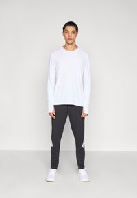 Biała sportowa koszulka z długim rękawem oraz ciemnoszare, zwężane spodnie z białymi paskami, do tego białe tenisówki, na neutralnym tle.