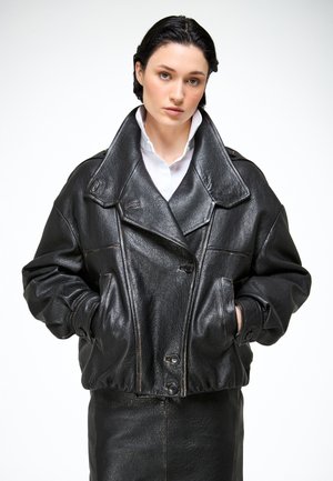 Femme aux cheveux courts et foncés portant une grande veste en cuir noire et une jupe assortie, les mains dans les poches, sur un fond blanc uni.