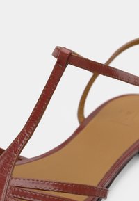 Κόκκινο σανδάλι από δερμάτινο βερνίκι με λεπτές διασταυρούμενες λουρίδες και επίπεδη, μπεζ σόλα. Ομαλή υφή με γυαλιστερό φινίρισμα.