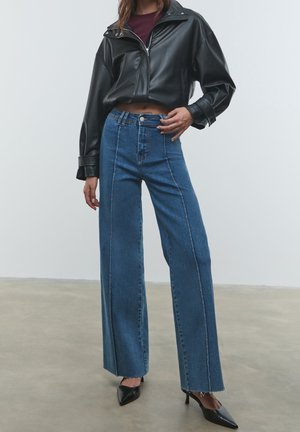 Femme portant une veste en cuir noire courte, un haut bordeaux, un jean large taille haute bleu, et des talons noirs à bout pointu, debout sur un sol gris.