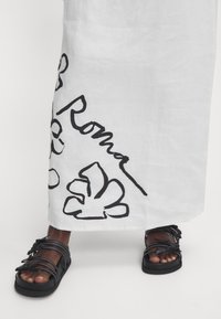 Mustat hihnaläpykkäät jaloissa, paksulla pohjalla, yhdistettynä valkoiseen hameeseen, jossa on rohkeita mustia kukka- ja tekstiprinttejä.