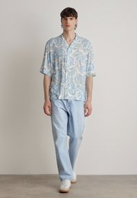 Maglietta azzurra a maniche corte con un motivo a vortice, abbinata a jeans in denim chiaro e sneakers bianche, in piedi contro uno sfondo neutro.