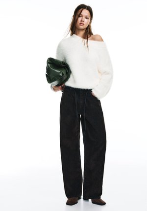 Ung kvinna står iklädd en vit off-shoulder fluffig tröja, lösa svarta byxor, bruna skor och håller en stor mörkgrön väska.