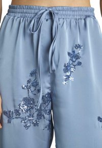 Lichtblauwe satijnen broek met een elastische tailleband en een trekkoord. Heeft donkerblauwe bloemenborduursels van pailletten aan één kant.