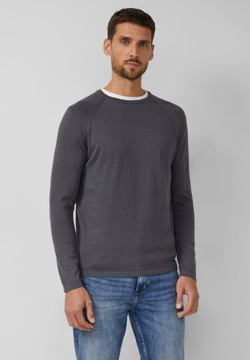 Mężczyzna stojący na szarym tle, ubrany w ciemnoszary longsleeve nałożony na biały t-shirt i niebieskie dżinsy.