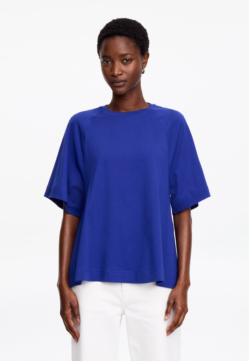 Femme portant un haut ample bleu roi à manches courtes et un pantalon blanc, debout devant un fond blanc uni.