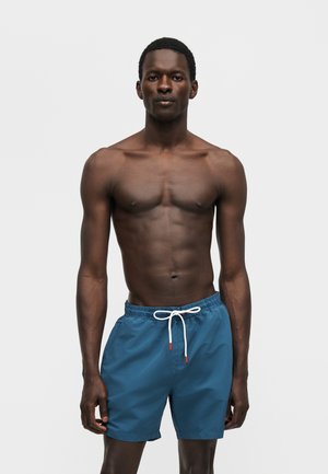 Ψηλός άνδρας με σκουρόχρωμο δέρμα όρθιος, φοράει μπλε μαγιό με λευκό κορδόνι, σε απλό ανοιχτό γκρι φόντο.