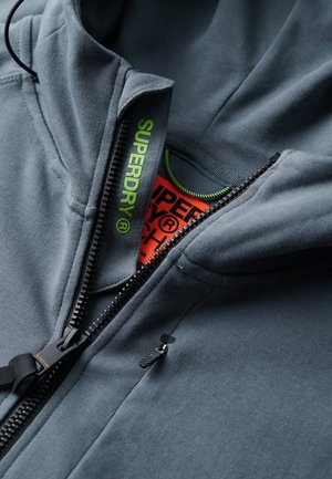 Grijze zip-up hoodie met een hoge kraag, zwarte rits, groene trekkoord en oranje label aan de binnenkant, voorzien van "SUPERDRY" merknaam in felgroen.