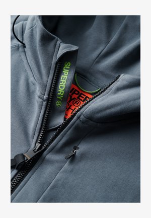 Grijze zip-up hoodie met een hoge kraag, zwarte rits, groene trekkoord en oranje label aan de binnenkant, voorzien van "SUPERDRY" merknaam in felgroen.