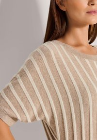 Vrouw in een beige trui met korte mouwen en witte verticale strepen, afgebeeld van schouder tot kin tegen een lichte achtergrond.