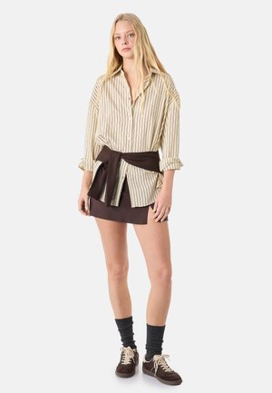 Jeune femme aux longs cheveux blonds porte une chemise rayée oversize, une mini-jupe marron, un pull foncé noué à la taille, des chaussettes grises et des baskets marron.