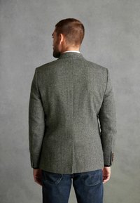 Blazer en pied-de-poule gris avec un design structuré, fente arrière unique et coupe ajustée, associé à un jean sombre. Détail de tissu texturé visible.