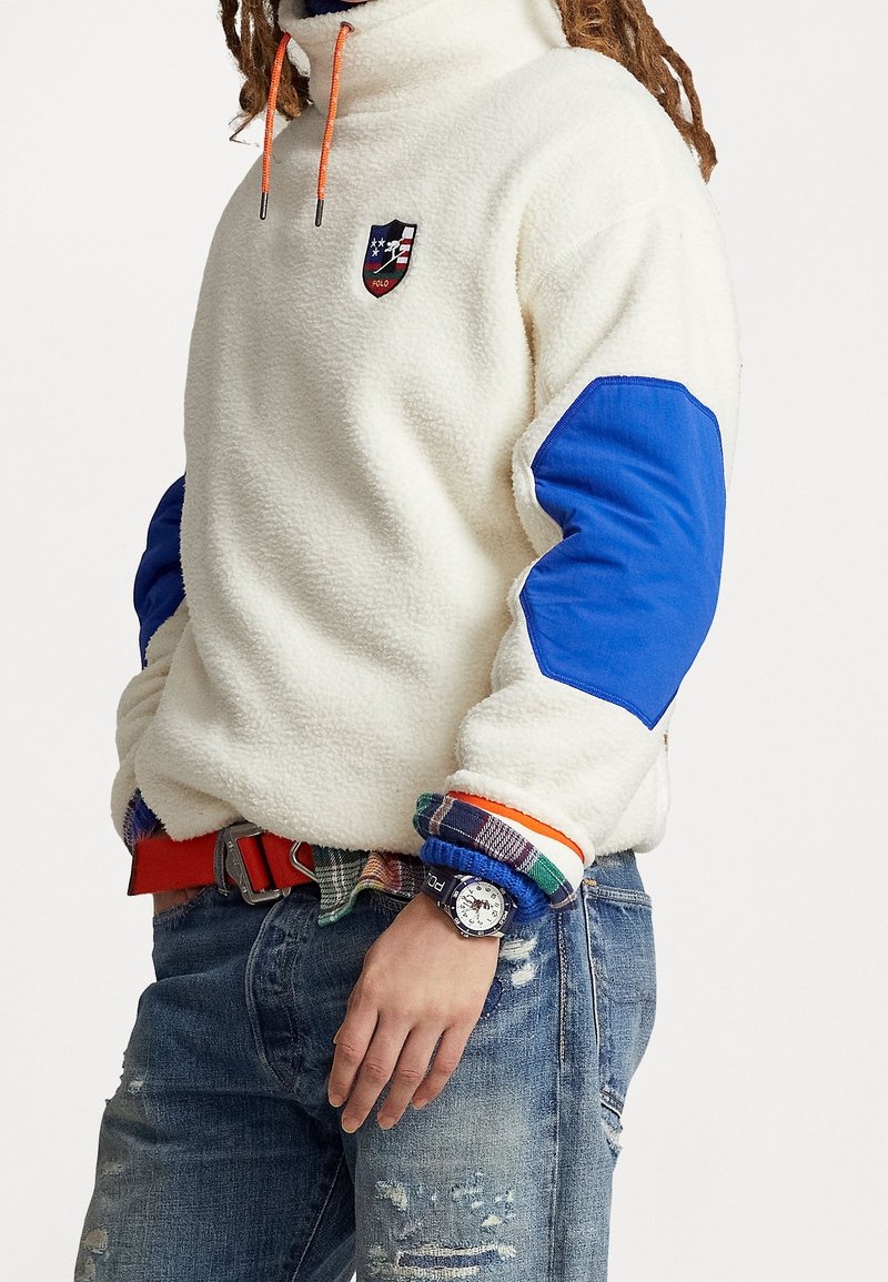 Fleece-pullover i elfenben med blå armbågsflickor, hög krage och orange dragband. Har ett logomärke och lager med rutiga skjortmuddar.