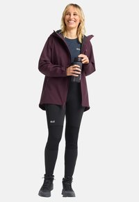Giacca impermeabile bordeaux con cappuccio, pantaloni termici neri e scarponi da trekking neri. Tiene in mano una bottiglia d'acqua nera, la texture è liscia e resistente.